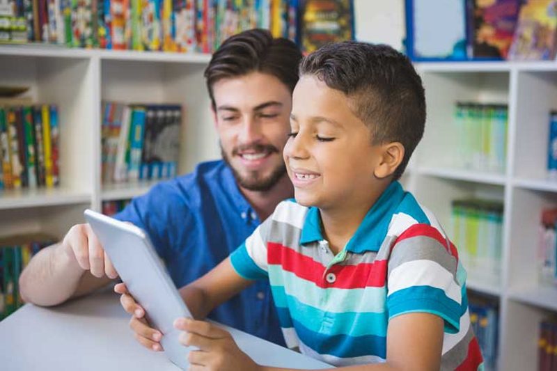 Elementary student smiling and holding tablet while teacher points at screen.