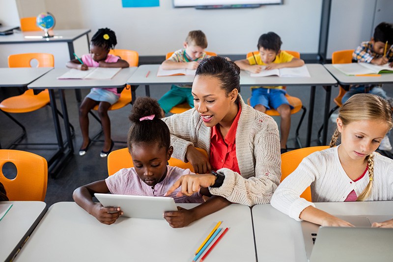 A young student looking at a tablet as their teacher points at it from over their shoulder