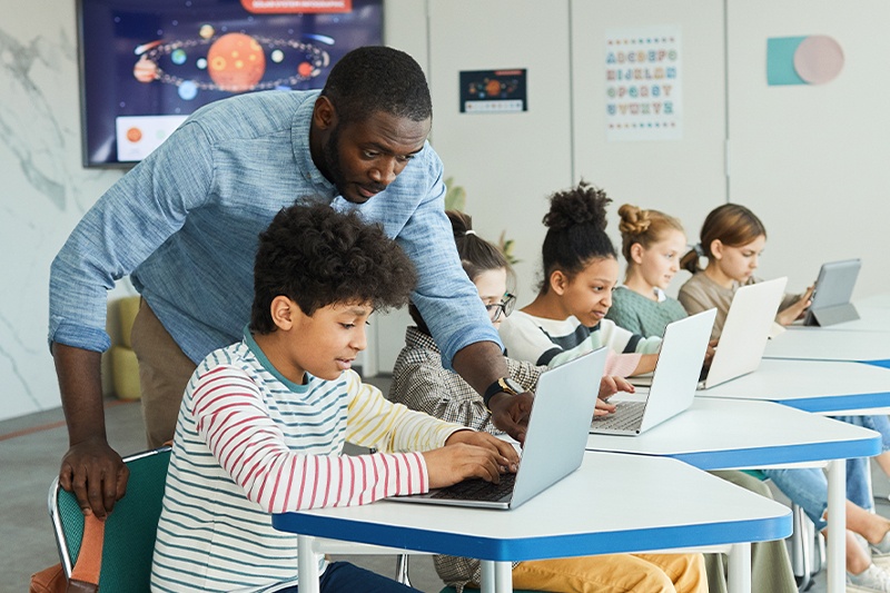 Middle school students working on laptops as a teacher observes and helps student