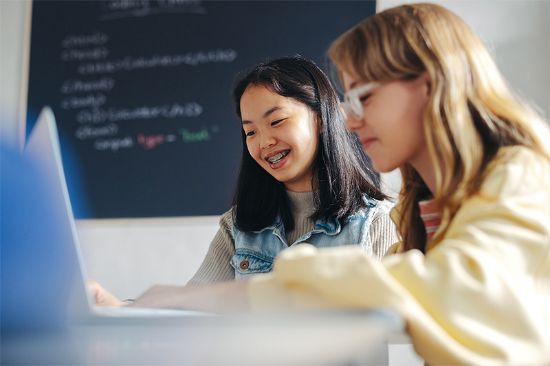 Two middle school students working on a laptop together with code shown on a display behind them.